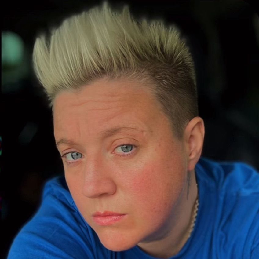 Close-up of a person with short, spiky blonde hair and blue eyes, wearing a blue shirt.