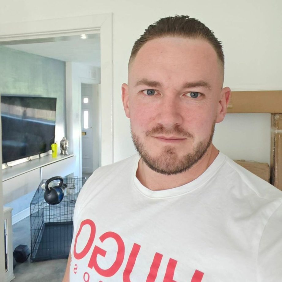 A man with short hair wearing a white T-shirt stands in a bright room.